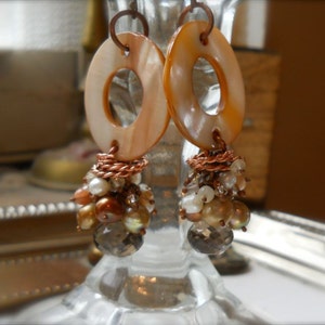 May include: A pair of handcrafted earrings featuring large, oval-shaped, light orange shell pendants with a central hole. Below, clusters of small, iridescent beads and faceted crystals dangle, connected by copper wire. The earrings are designed for pierced ears.