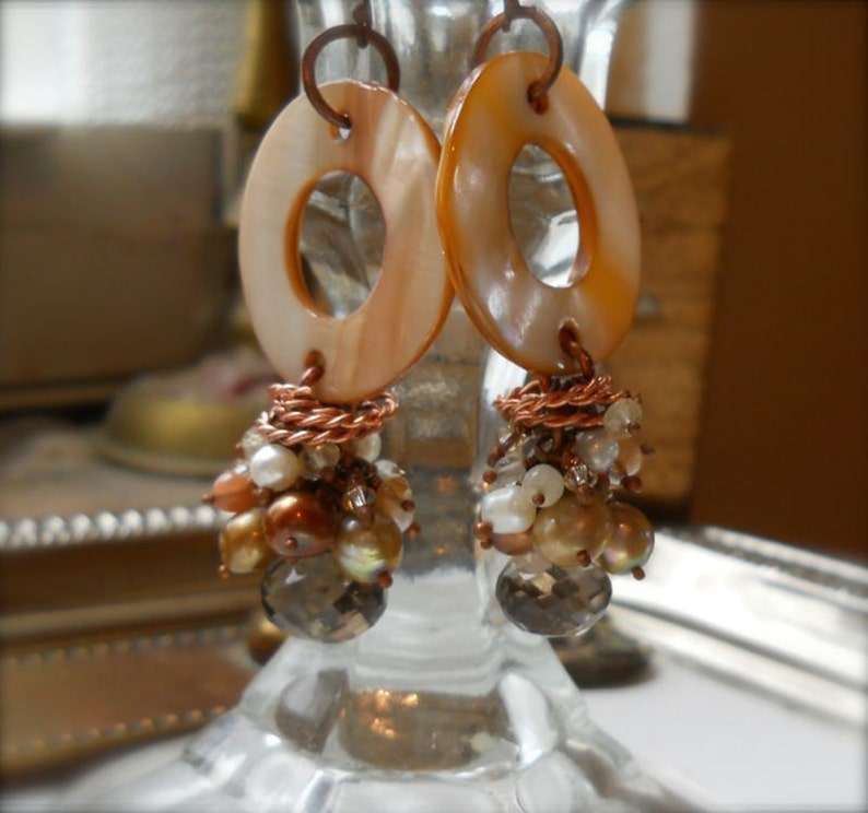May include: A pair of handcrafted earrings featuring large, oval-shaped, light orange shell pendants with a central hole. Below, clusters of small, iridescent beads and faceted crystals dangle, connected by copper wire. The earrings are designed for pierced ears.