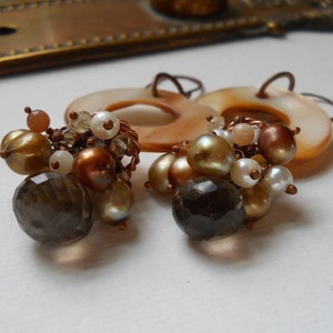 May include: A pair of handcrafted earrings featuring a cluster of beads in shades of gold, brown, and white. The earrings include faceted smoky quartz, pearl-like beads, and small round beads, all connected with copper wire. The earrings have a circular, shell-like backing.