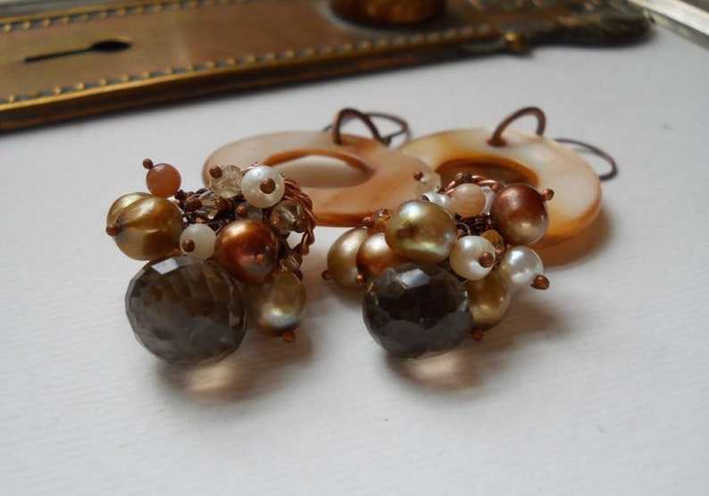 May include: A pair of handcrafted earrings featuring a cluster of beads in shades of gold, brown, and white. The earrings include faceted smoky quartz, pearl-like beads, and small round beads, all connected with copper wire. The earrings have a circular, shell-like backing.