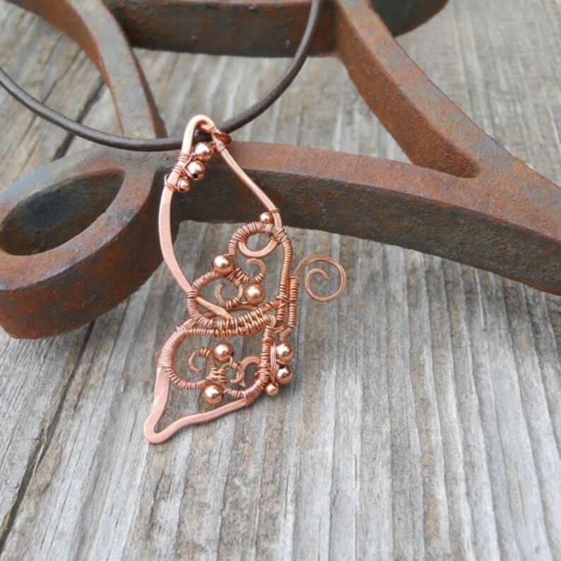 May include: Copper wire pendant with a butterfly wing design. The pendant features intricate wire wrapping with small copper beads and a swirling detail. It hangs from a dark brown cord, set against a rustic wooden surface.