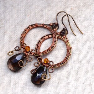 May include: Handmade copper wire earrings with a circular design. Each earring features a teardrop-shaped, dark brown gemstone and small amber-colored beads. The earrings have a rustic, bohemian style.