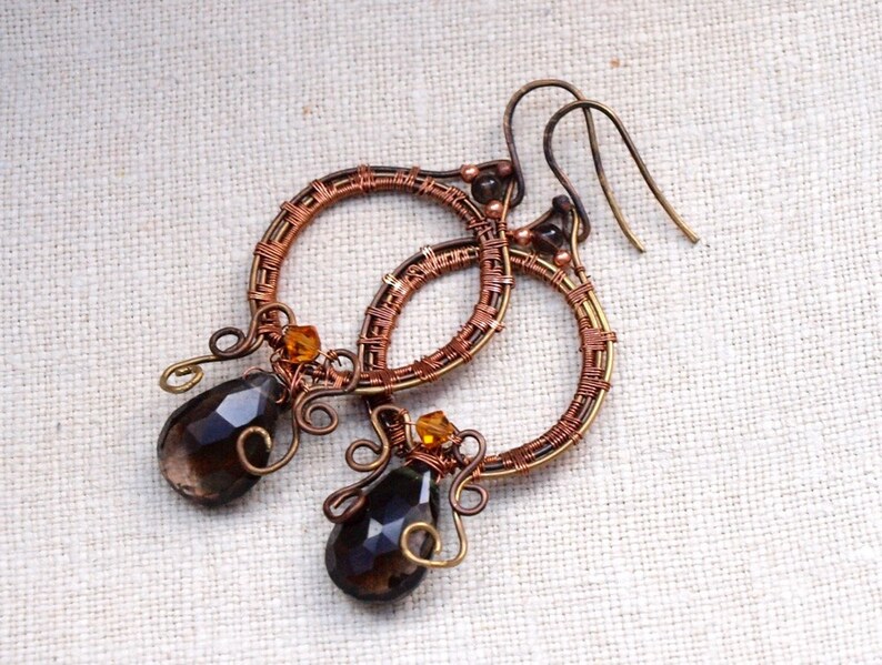 May include: Handmade copper wire earrings with a circular design. Each earring features a teardrop-shaped, dark brown gemstone and small amber-colored beads. The earrings have a rustic, bohemian style.