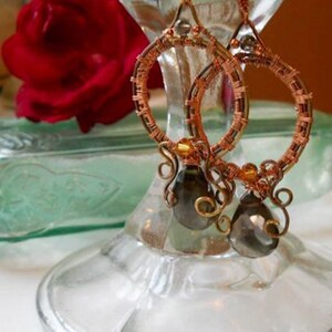 May include: Handmade copper wire hoop earrings with smoky gray teardrop beads. The hoops are intricately wrapped with copper wire, featuring decorative swirls and gold-colored accents. The earrings are displayed on a glass stand, with a red rose and a vintage green glass bottle in the background.