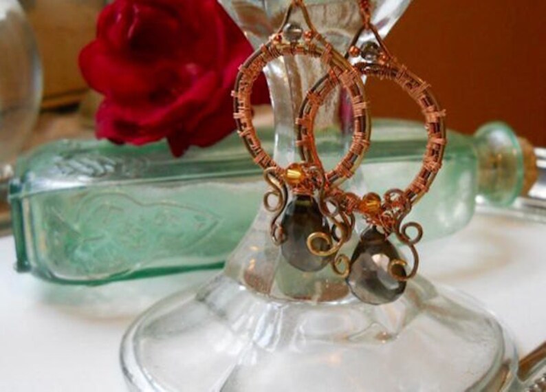 May include: Handmade copper wire hoop earrings with smoky gray teardrop beads. The hoops are intricately wrapped with copper wire, featuring decorative swirls and gold-colored accents. The earrings are displayed on a glass stand, with a red rose and a vintage green glass bottle in the background.