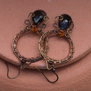 May include: Handmade copper wire earrings with a unique design. Each earring features a dark brown, faceted gemstone and a smaller orange bead. The hoops are intricately wrapped with copper wire, creating a textured effect. The earrings have a hook closure.