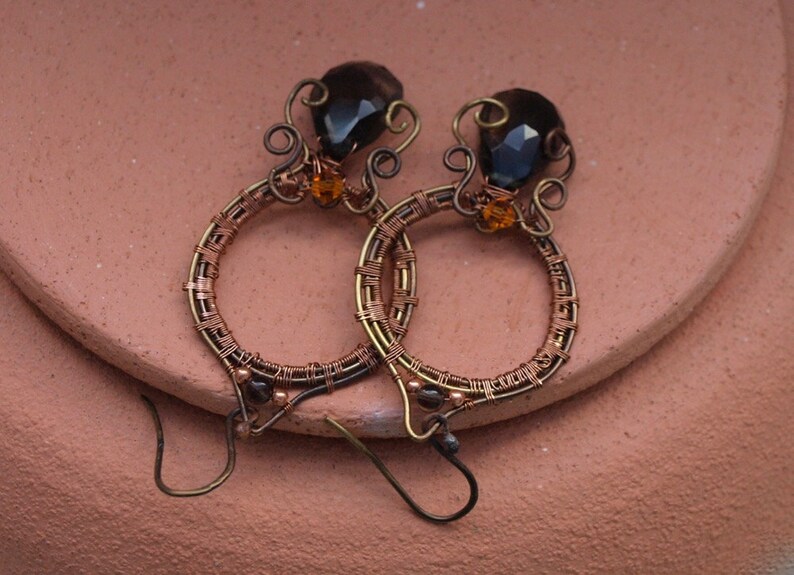 May include: Handmade copper wire earrings with a unique design. Each earring features a dark brown, faceted gemstone and a smaller orange bead. The hoops are intricately wrapped with copper wire, creating a textured effect. The earrings have a hook closure.