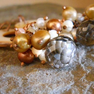 May include: A close-up of a bracelet with a mix of beads in various colors, including gold, bronze, white, and a faceted gray crystal. The bracelet is set against a textured background.
