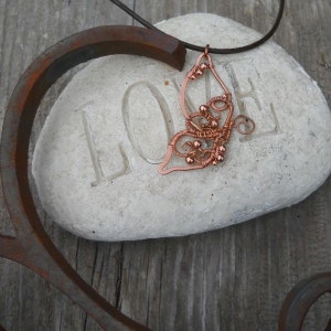 May include: A copper-colored wire-wrapped butterfly pendant on a leather cord. The pendant is resting on a white stone with the word "LOVE" carved into it. The butterfly design features intricate wire detailing and small copper beads.