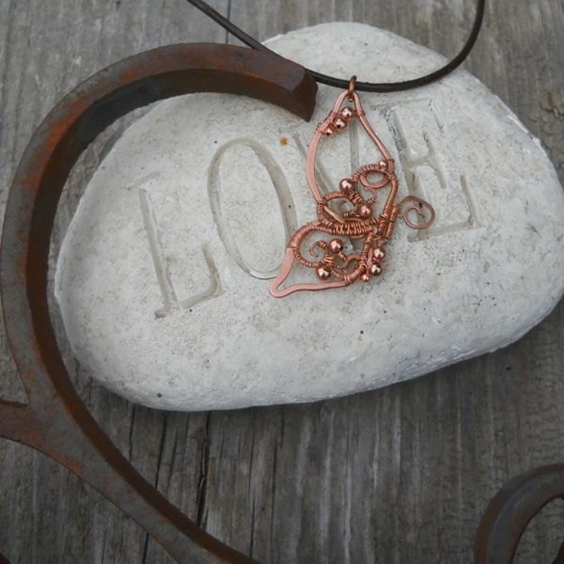 May include: A copper-colored wire-wrapped butterfly pendant on a leather cord. The pendant is resting on a white stone with the word "LOVE" carved into it. The butterfly design features intricate wire detailing and small copper beads.
