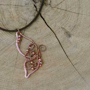 May include: A copper-colored wire pendant necklace in the shape of a butterfly wing. The pendant features intricate wire wrapping and small copper beads. The necklace hangs on a dark brown leather cord. The background is a light brown wooden surface.