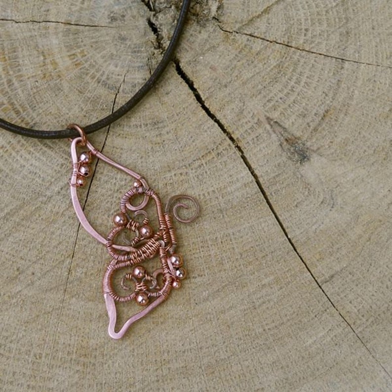 May include: A copper-colored wire pendant necklace in the shape of a butterfly wing. The pendant features intricate wire wrapping and small copper beads. The necklace hangs on a dark brown leather cord. The background is a light brown wooden surface.