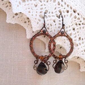 May include: Handmade copper wire hoop earrings with dark brown, teardrop-shaped beads. The earrings feature intricate wire wrapping and small, amber-colored beads. The hoops are suspended from a simple hook, showcasing a bohemian style.