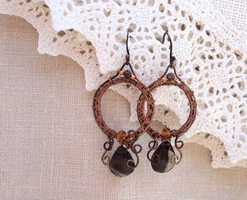 May include: Handmade copper wire hoop earrings with dark brown, teardrop-shaped beads. The earrings feature intricate wire wrapping and small, amber-colored beads. The hoops are suspended from a simple hook, showcasing a bohemian style.