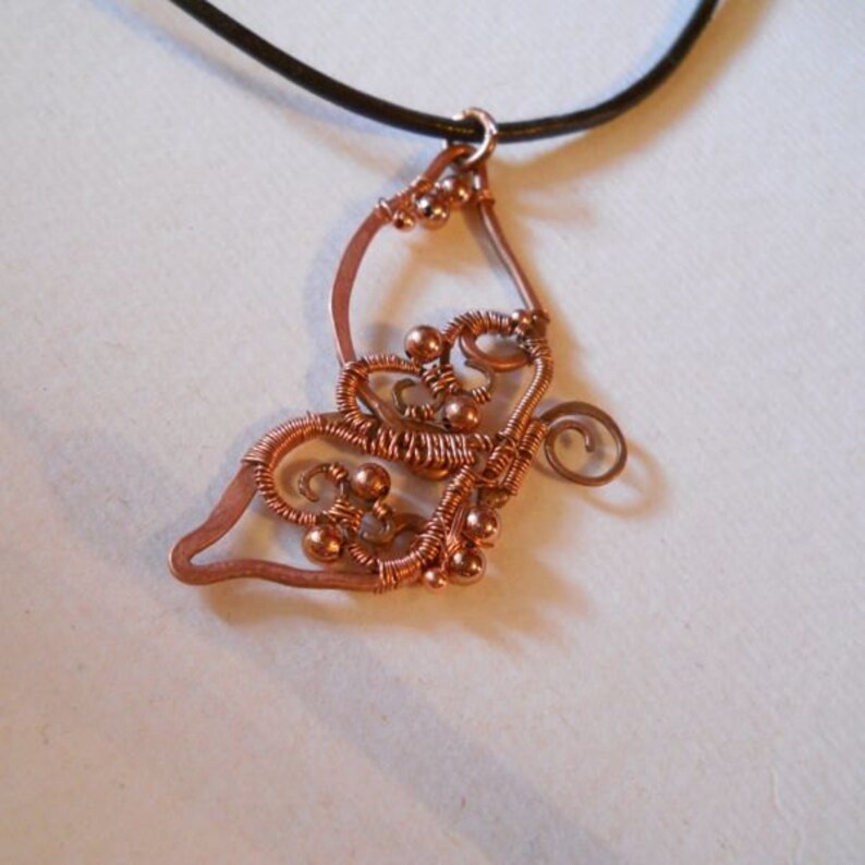 May include: Copper wire pendant with a butterfly design. The pendant features intricate wire wrapping and small copper beads. It hangs from a black leather cord, creating a unique and artistic necklace.