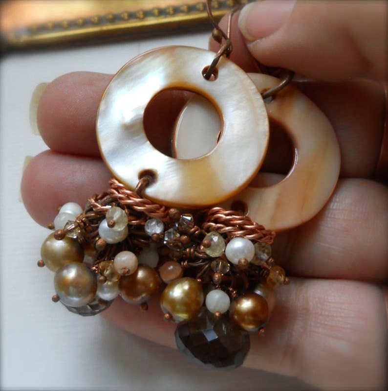 May include: Handmade earrings featuring large, circular, iridescent mother-of-pearl pendants. Below, a cluster of pearls, beads, and crystals in shades of gold, brown, and white are attached with copper wire. The earrings are held up by copper hooks.