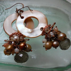 May include: A pair of dangle earrings featuring large, round, iridescent mother-of-pearl discs. The earrings have clusters of brown, gold, and white beads, with a faceted smoky quartz bead. The earrings are connected with copper wire.
