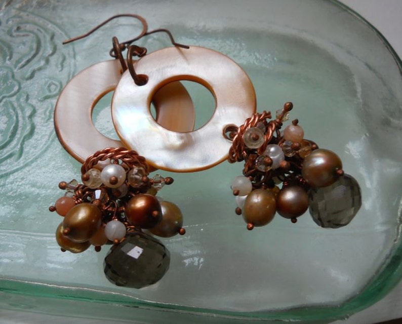May include: A pair of dangle earrings featuring large, round, iridescent mother-of-pearl discs. The earrings have clusters of brown, gold, and white beads, with a faceted smoky quartz bead. The earrings are connected with copper wire.