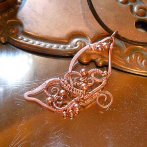 May include: Copper wire pendant with intricate woven and coiled design. The pendant features a teardrop shape with decorative swirls and small bead accents. The warm copper color contrasts with the reflective surface it rests on.