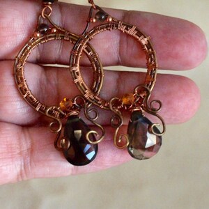 May include: Handmade copper wire hoop earrings with brown and amber-colored accents. The earrings feature a teardrop-shaped, faceted gemstone pendant. The hoops are intricately wrapped with copper wire, creating a unique, bohemian style.