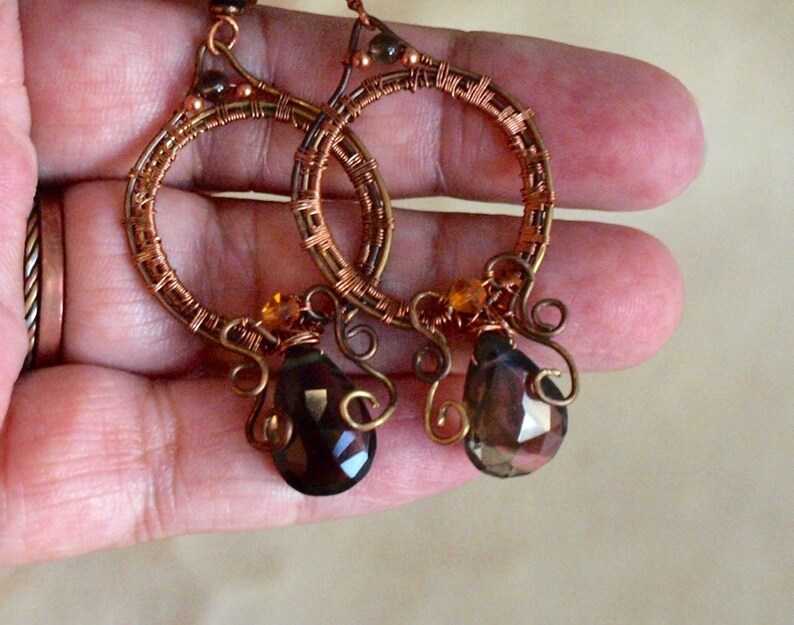 May include: Handmade copper wire hoop earrings with brown and amber-colored accents. The earrings feature a teardrop-shaped, faceted gemstone pendant. The hoops are intricately wrapped with copper wire, creating a unique, bohemian style.