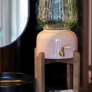 Pode incluir: Um dispensador de água de cerâmica rosa com uma torneira dourada, encimado por um recipiente de vidro verde. Ele está em um suporte de madeira. Dois copos transparentes estampados estão na frente. Um dispensador de água elegante.