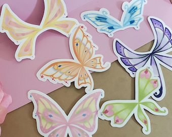 Puede incluir: Seis pegatinas de mariposas de colores con diferentes formas y patrones de alas. Las pegatinas están sobre un fondo rosa.