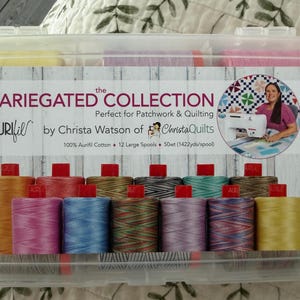 May include: A clear plastic storage case containing spools of Aurifil variegated cotton thread. The case features a label with text that reads "The Variegated Collection" and "Perfect for Patchwork & Quilting". The spools are in various colors.