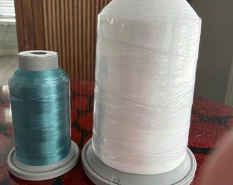 May include: Two spools of thread standing upright. The smaller spool on the left is a metallic teal color, while the larger spool on the right is white. Both spools feature a gray plastic base and a gray top cap, with the thread wound tightly.