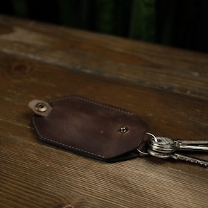 Puede incluir: Un llavero de cuero marrón con un borde cosido y dos broches metálicos. El llavero está sobre una superficie de madera con un juego de llaves adjunto.