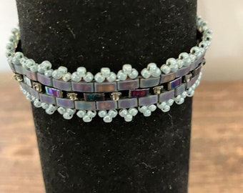 May include: A beaded bracelet with a silver and iridescent blue design. The bracelet is on a black display stand.