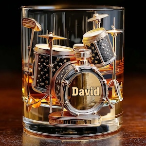 Puede incluir: Un vaso transparente con un líquido dorado, que contiene una intrincada escultura de batería en tono dorado en su interior. El bombo se muestra prominentemente en el centro con el nombre David grabado en letras oscuras y negritas al frente.