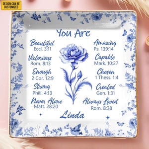 Può includere: Un piattino quadrato in ceramica bianca con bordo floreale blu e un'illustrazione di un fiore blu al centro. Il testo recita: You Are Beautiful, Amazing, Victorious, Capable, Enough, Chosen, Strong, Created, Never Alone, Always Loved, Linda.