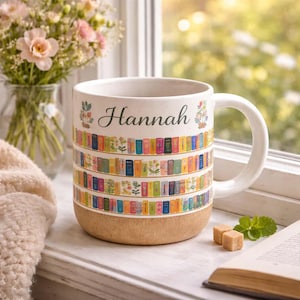 May include: A white ceramic mug with the name "Hannah" in script, decorated with colorful rows of books. The bottom of the mug has a textured, tan finish. The mug is on a windowsill with a book, mint, and sugar cubes.