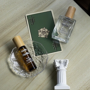 May include: Two glass bottles of Nuvia No. 12 Petal Drift perfume, one in a rollerball and one in a spray bottle, on a wooden surface with a green card and a white decorative column.
