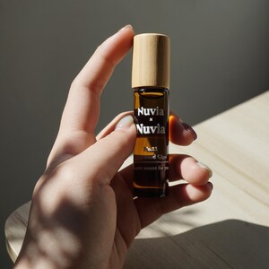 May include: A brown glass roller bottle with a wooden cap. The bottle is labelled "Nuvia x Nuvia No.11 Rosewood Glow scent meant for you."
