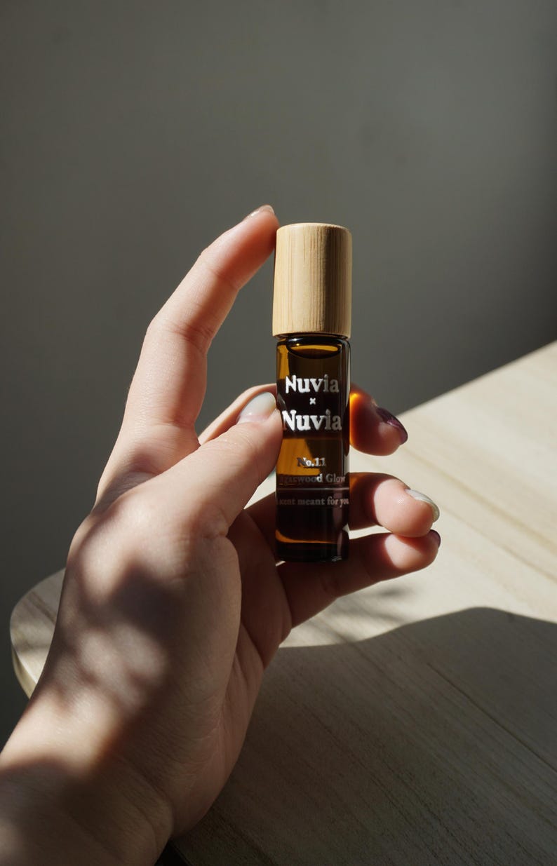 May include: A brown glass roller bottle with a wooden cap. The bottle is labelled "Nuvia x Nuvia No.11 Rosewood Glow scent meant for you."