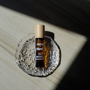 May include: A brown glass roller bottle with a wooden cap and a white label that reads "Nuvia x Nuvia No.11 Agarwood Oil Scent meant to..." The bottle is sitting on a clear glass dish with a textured surface.