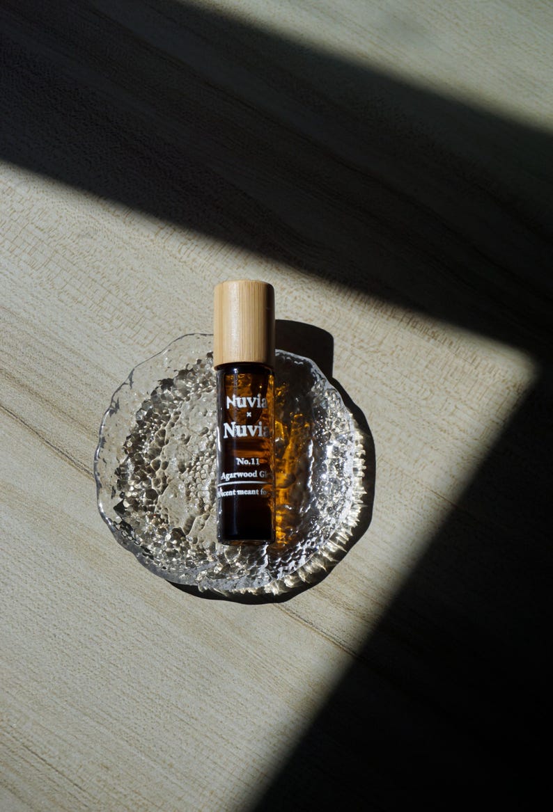 May include: A brown glass roller bottle with a wooden cap and a white label that reads "Nuvia x Nuvia No.11 Agarwood Oil Scent meant to..." The bottle is sitting on a clear glass dish with a textured surface.