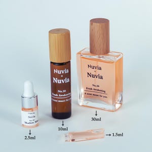 May include: Three glass bottles of perfume with wooden caps. The bottles are labelled "Nuvia x Nuvia" and "Nuvia Nuvia" with the text "No. 20 Fresh Awakening A scent meant for you." The bottles are 2.5 ml, 10 ml, and 30 ml.