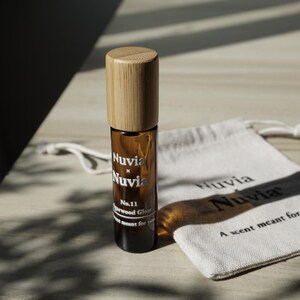 May include: A brown glass roll-on bottle with a bamboo lid and a white label that reads "Nuvia x Nuvia No. 11 Sandalwood Glow A scent meant for you." The bottle is sitting on a white fabric pouch with the text "Nuvia Nuvia A scent meant for you."