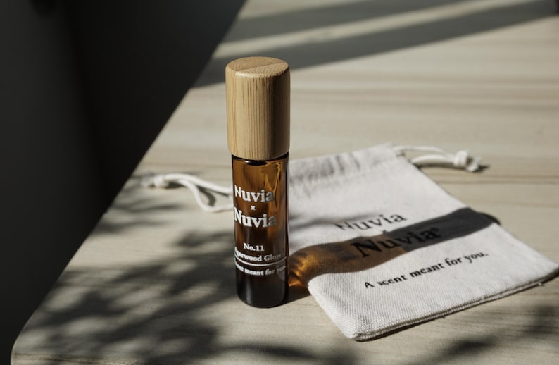 May include: A brown glass roll-on bottle with a bamboo lid and a white label that reads "Nuvia x Nuvia No. 11 Sandalwood Glow A scent meant for you." The bottle is sitting on a white fabric pouch with the text "Nuvia Nuvia A scent meant for you."