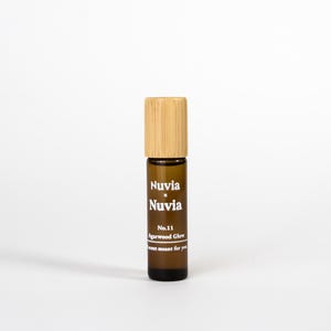 May include: A brown glass roller bottle with a bamboo lid. The bottle is labelled with the text "Nuvia x Nuvia", "No. 11 Agarwood Glow", and "Just meant for you".