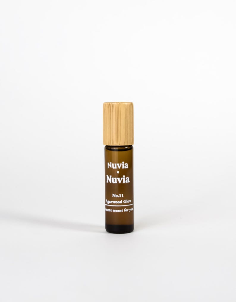 May include: A brown glass roller bottle with a bamboo lid. The bottle is labelled with the text "Nuvia x Nuvia", "No. 11 Agarwood Glow", and "Just meant for you".