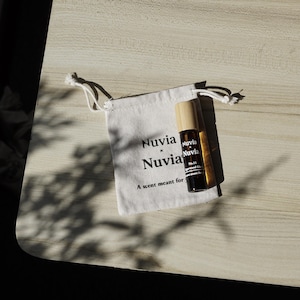 Puede incluir: Una bolsa blanca de cordón con el texto "Nuvia x Nuvia" y la frase "A scent meant for..." impresa en ella. Una pequeña botella de vidrio marrón con una tapa de madera y el texto "Nuvia" está dentro de la bolsa.