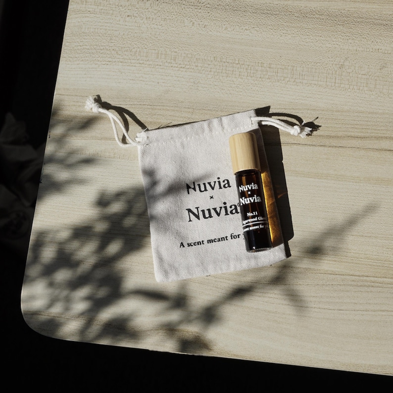 May include: A white drawstring bag with the text "Nuvia x Nuvia" and the phrase "A scent meant for..." printed on it. A small brown glass bottle with a wooden cap and the text "Nuvia" is inside the bag.