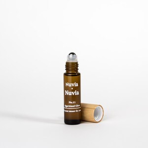 May include: A brown glass roller bottle with a silver roller ball. The bottle is labelled "Nuvia x Nuvia No. 11 Agarwood Glow A scent meant for you". The bottle is sitting on a white surface next to a bamboo cap.