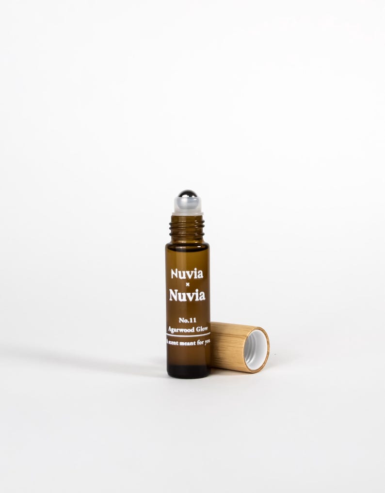 May include: A brown glass roller bottle with a silver roller ball. The bottle is labelled "Nuvia x Nuvia No. 11 Agarwood Glow A scent meant for you". The bottle is sitting on a white surface next to a bamboo cap.