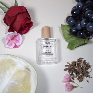Puede incluir: Un frasco de perfume de vidrio transparente con tapón de madera, etiquetado "Nuvia" y "Cedar Bloom". La imagen incluye una rosa roja, flores rosas, uvas y una rodaja de cítricos, lo que sugiere una fragancia floral y afrutada.