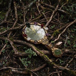 Op de afbeelding: Een goudkleurige handspiegel met een ronde spiegel en een handvat. De spiegel is versierd met kleurrijke edelstenen en een pentagramsymbool. Het handvat heeft aan het uiteinde een groene edelsteen. De spiegel ligt op een achtergrond van boomwortels en groen.